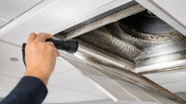 Technician using a flashlight to inspect a commercial air duct vent for dust buildup