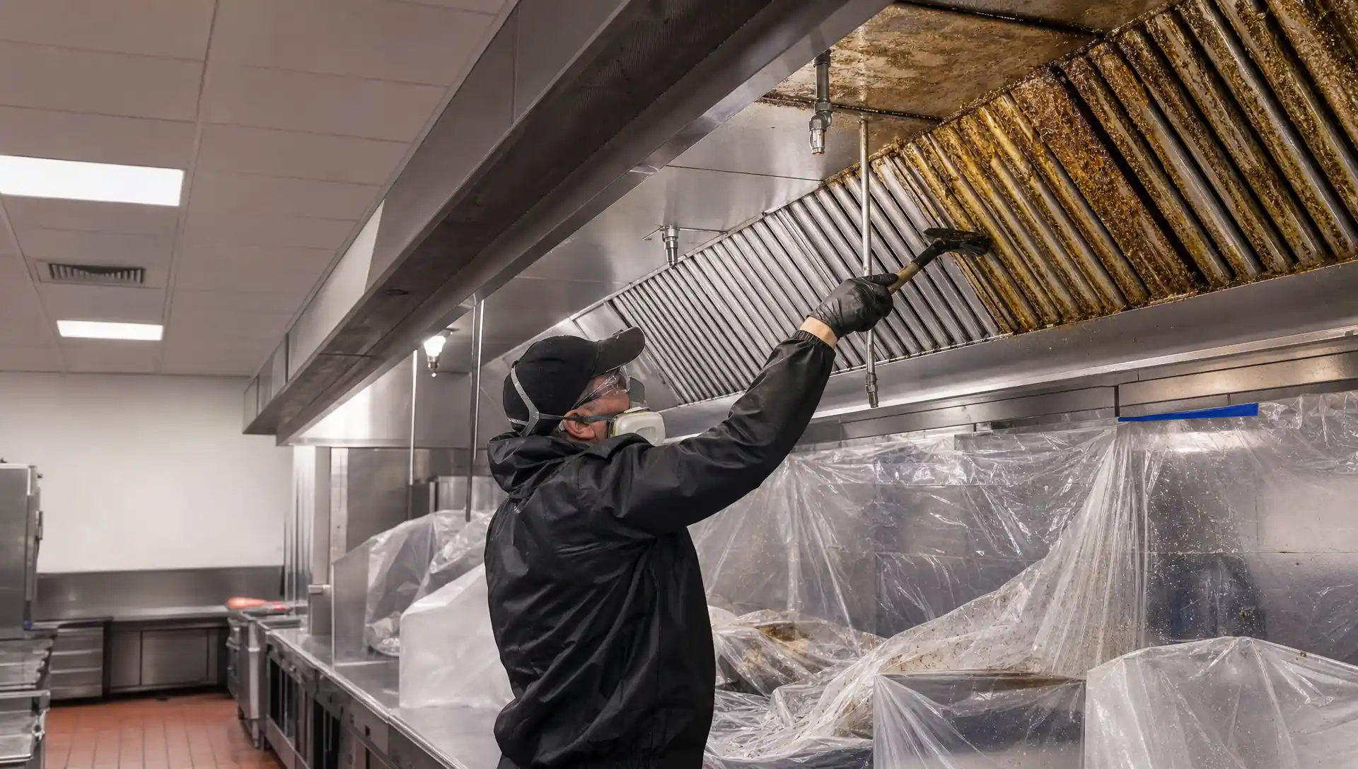 Service-Tech technician cleaning a commercial kitchen exhaust hood and grease duct to bare metal per NFPA 96 with cookline protection in place